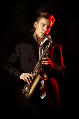 Fototapeta na wymiar Young guy posing in a stylish black suit with a saxophone in his hands and red illumination