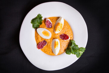 Vegetable cream soup with egg, salami slices, and cilantro on a white plate on black background.