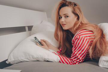 A beautiful girl on a white bed in red striped pajamas cannot sleep, looks at the phone. Sleeplessness, a smartphone in the hands of a woman since the morning. Social addiction. Works in bed.
