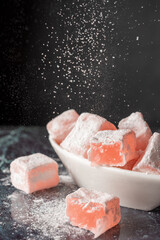 Fototapeta premium Top view of Turkish Delight with roses in white bowl, with sugar falling on blue marble, selective focus and black background, vertical