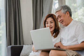 Asian senior couple using laptop at home. Asian Senior Chinese grandparents, surf the Internet to check social media while lying on sofa in living room at home concept.