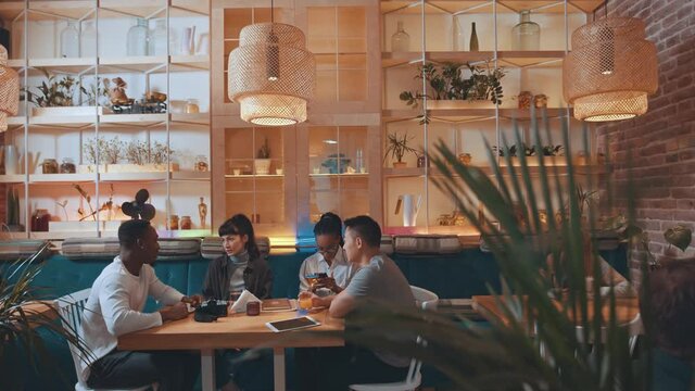 Multicultural happy friends talking and laughing while sitting at the cafeteria table together indoors. Smiling diverse young mates students having fun and drinking wine together at meeting at table