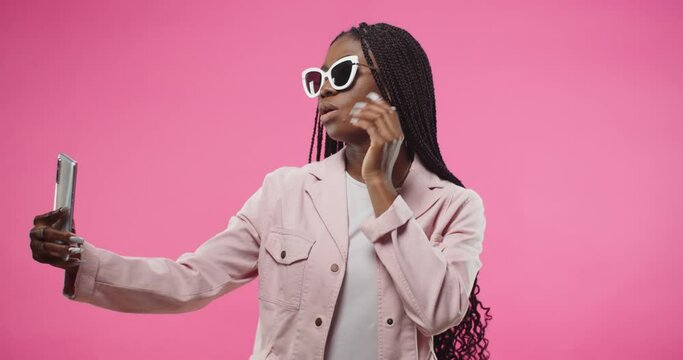 Portrait of happy cheerful young beautiful African American woman in pink jacket and sunglasses posing isolated on rosy background taking selfie photos on smartphone, photoshoot on cellphone