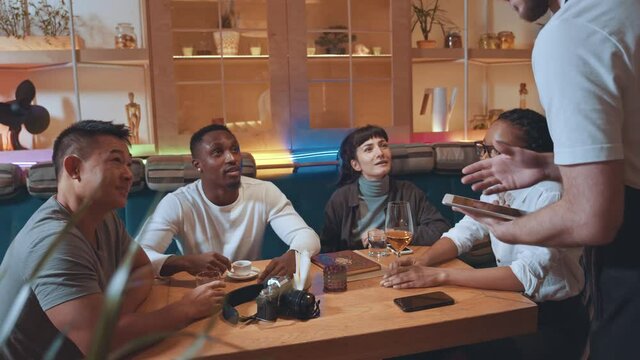 High angle view of handsome young multiracial company of friends talking to waiter for ordering drinks in beautiful modern cafe interior. People chatting with bartender. Customers at restaurant