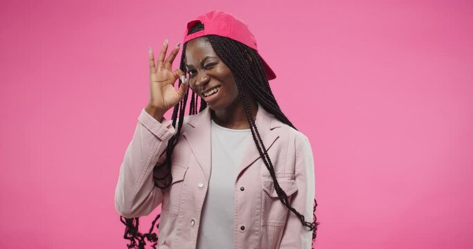 Portrait of African American young pretty cheerful woman wearing red cap in positive mood smiling and making OK gestures with fingers approving with hand posing isolated on rosy background in studio