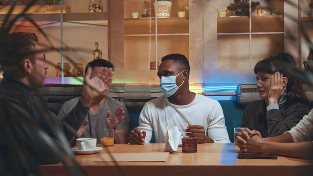 Happy diverse people persuade their friend to take off his protective mask while he sitting at the cafeteria. Friends having fun together and do not comply with the quarantine rules. Pandemic concept