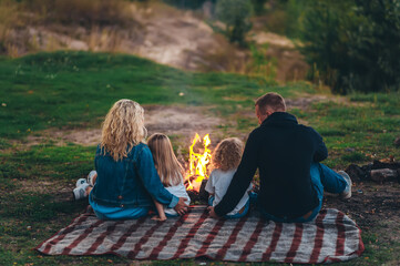 Happy family of four is sitting on the green grass near campfire.
