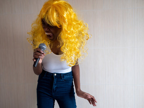 African american woman in white t-shirt in bright yellow wig sings karaoke with microphone in her hands on light background, posing for camera