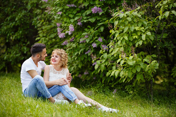 Naklejka na ściany i meble Young man and woman couple near a blooming lilac bush. Tender holding each other. Spring lovestory. Blonde-haired girl with curled hairs and man weared in casual. Young family