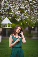 Fototapeta premium Young woman in long green dress in a blooming apple garden. Spring story. Brown-haired girl with long hairs.
