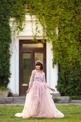 Obraz premium Beautiful woman in flying airy pink dress on background of door overgrown with wild grapes. Pretty young caucasian girl plays with her dress. Girl dance in flowing dress