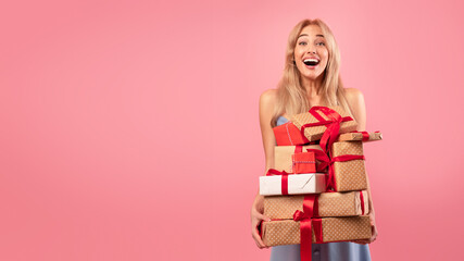 Obraz na płótnie Canvas Young blonde woman with pile of wrapped gift boxes looking at camera and smiling in excitement on pink studio background