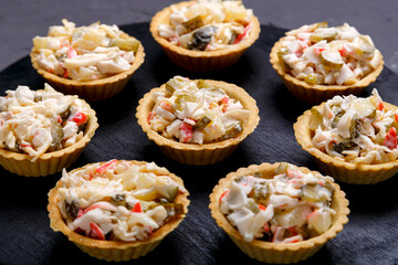 Tartlets with salad on a black wooden round board laid out in a circle on a gray concrete background.