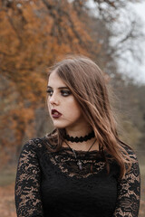Portrait of a young witch on a background of a gloomy autumn garden. The girl is wearing a black lace dress. The gaze is set aside. On the face - dark gothic makeup. Shooting for Halloween.
