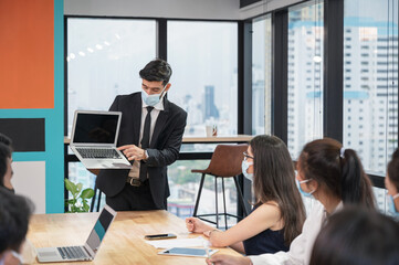 Businessman wearing face mask presentation of business plan on laptop in multiethnic business meeting at modern office