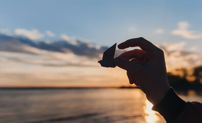 A female hand holds a paper crane against the backdrop of a sunset and sky with clouds. Origami, bird. The Japanese concept of peace and happiness.
