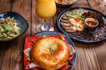 Serving various dishes with sliced vegetables, meat cuts, salads, orange juice and a bun on a brown wooden background.