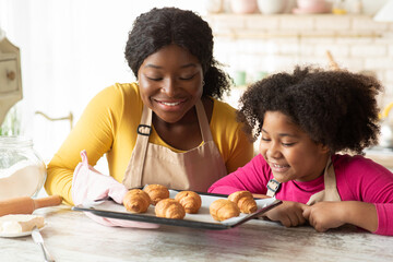 Black mother and daughter holding tray with fresh baked croissants in kitchen - obrazy, fototapety, plakaty