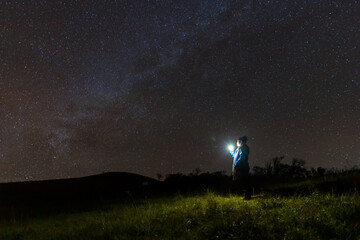 Naklejka na ściany i meble Climber or backpacker. Light holds up over his head. Standing on mountain peak. man with flashlight point to the starry sky at midnight. tourist with flashlight