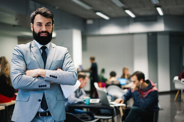 Cheerful businessman standing with crossed arms near colleagues working in modern office