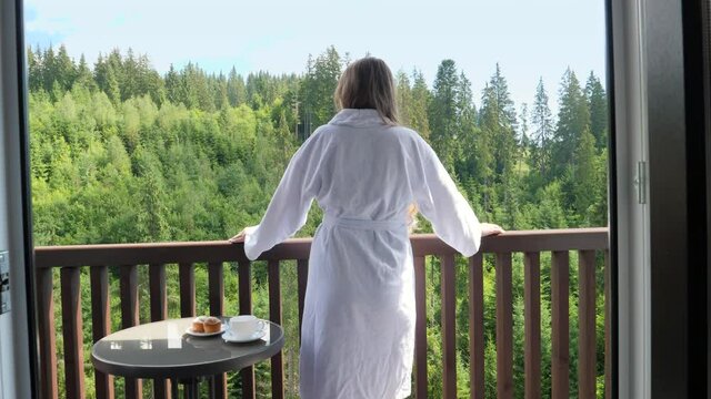 Beautiful young woman in bathrobe standing on hotel terrace or balcony and looking on the pine forest and mountains. Concept of tourism and vacation in mountains at summer