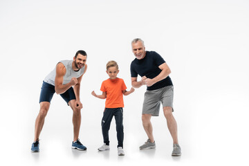Obraz na płótnie Canvas boy with dad and grandfather in sportswear demonstrating strength while smiling at camera on white