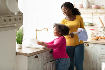Little Helper. Cute Black Preschool Girl Washing Dishes With Mom In Kitchen