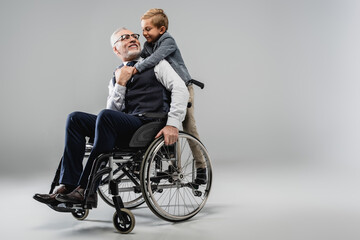 happy man in wheelchair looking at smiling grandson hugging him on grey