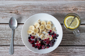 Oatmeal with banana pieces and berries, drenched in milk. Next to a glass of water with lemon