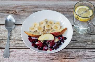 Oatmeal with banana pieces and berries, drenched in milk. Next to a glass of water with lemon