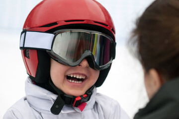Cheerful boy in red helmet, ski goggles and white jacket smiling to his mother. Winter sports, young skier