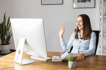 Cheerful female office employee in smart casual wear waving into webcam, talking online in virtual...