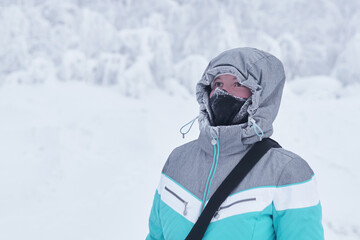 Fototapeta na wymiar young woman in a warm jacket with a hood and a buff neck warmer on her face against the background of blurred winter landscape