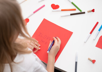 The child draws a postcard for the holiday of Mother's Day. Girl writes on red paper I love you mom