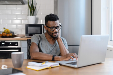 Tired african american male employee sit at the table in home office, using laptop, smiling....