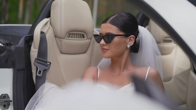 Stylish bride in sunglasses sits in a convertible. A bride in a white dress is sitting in the car. Beauty bride in wedding dress in the car. A dark-haired bride, wearing sunglasses, sits in a black ca