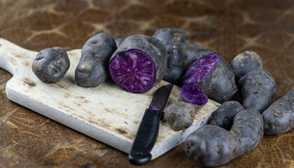 Vitelotte, french purple potatoes sliced in half with a knife on wooden board at rustic kitchen table. Blue variety of popular potato