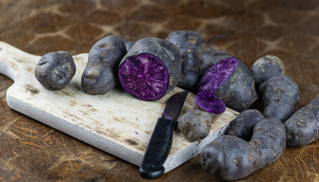 Vitelotte, french purple potatoes sliced in half with a knife on wooden board at rustic kitchen table. Blue variety of popular potato