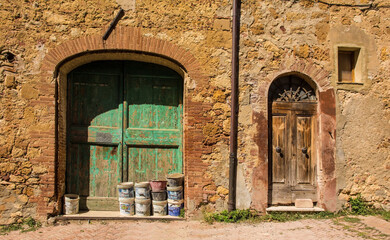 Fototapeta na wymiar Old wooden doors in an historic stone residential building in the village of Pienza in Siena Province, Tuscany, Italy. The green door has a variety of paints, building materials and buckets on its ste