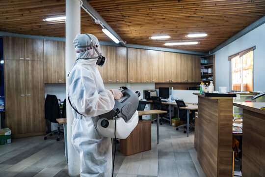 Professional technical man in prevention suit with his sterilizing machine and disinfecting water sprays all areas in the office room to take purifying coronavirus (COVID-19).