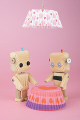 a pair of wooden robots near a toy table made from a cupcake pan with a lamp shining from above. St. Valentine's Day