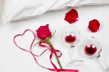 Fototapeta na wymiar Red heart of ribbon with red rose, rose petals and wine glasses with red wine on white bed honeymoon. Surprise Valentine and wedding day in bed. Top view. Copy space