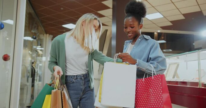 Two multi-cultural stylish women walk into shopping mall on sale boasting their purchases enjoying weekend together. Friendship. Shopaholic lifestyle.