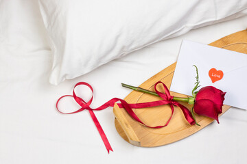 Red rose and envelope with little heart and ribbon on white bed with pillow, Valentin day flat lay top view romantic concept background.