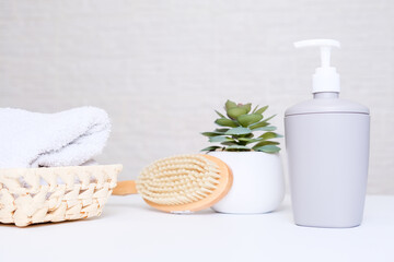 Fototapeta na wymiar Grey soap dispenser, spa towel, plant and massage brush on white counter table inside a bright bathroom background