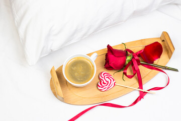 Romantic Valentine’s Day or holiday breakfast in bed with cup of coffee, heart shape lollipop and red rose. Top view, flat lay image.