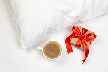 Fototapeta na wymiar Cup of coffee and gift box with red ribbon on white bed with pillow. Morning romantic surprise flat lay background.