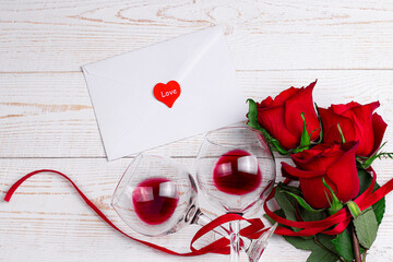 Wine glasses with red wine, envelope and red roses with red ribbon on white wooden background. Valentine day romantic dinner flat lay top view concept with copy space.