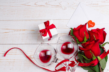 Wine glasses with red wine, gift box, envelope and red roses with red ribbon on white wooden background. Valentine day romantic dinner flat lay top view concept.