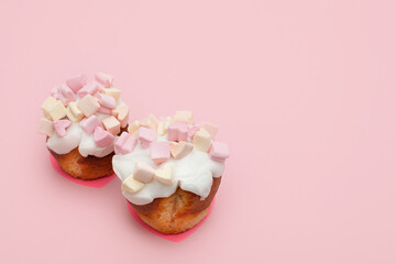 two freshly baked cupcakes covered with sweet icing and heart-shaped marshmallows on a pink background